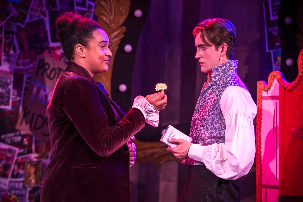 Two actors face each other, one in a purple jacket, smiling, offering a green carnation to another. 