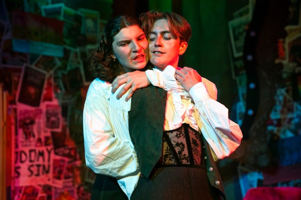 One actor is stood behind another, with his arm wrapped around the others upper chest, holding him tight. 