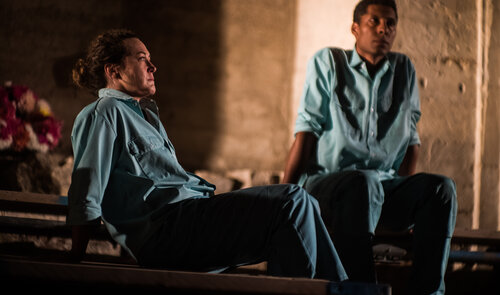 Two actors sit wearing hospital uniforms.