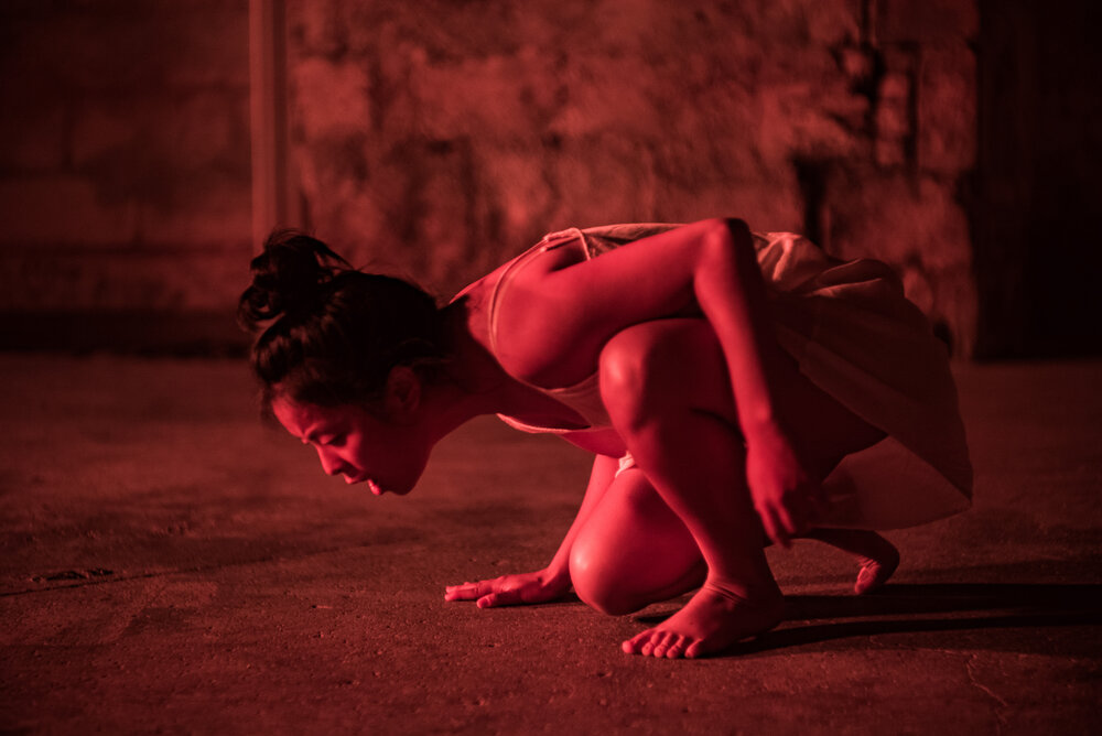 A dancer bathed in red light is crouched on the ground.
