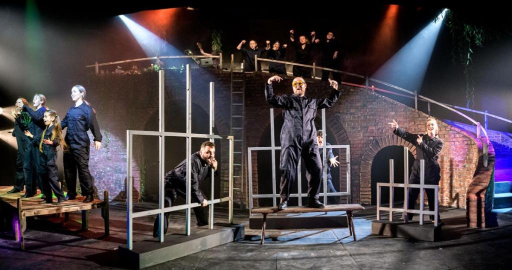 Three wooden frames in the shape of prison bars are in front of three actors in jumpsuits. In the foreground is another actor stood dancing on a bench.