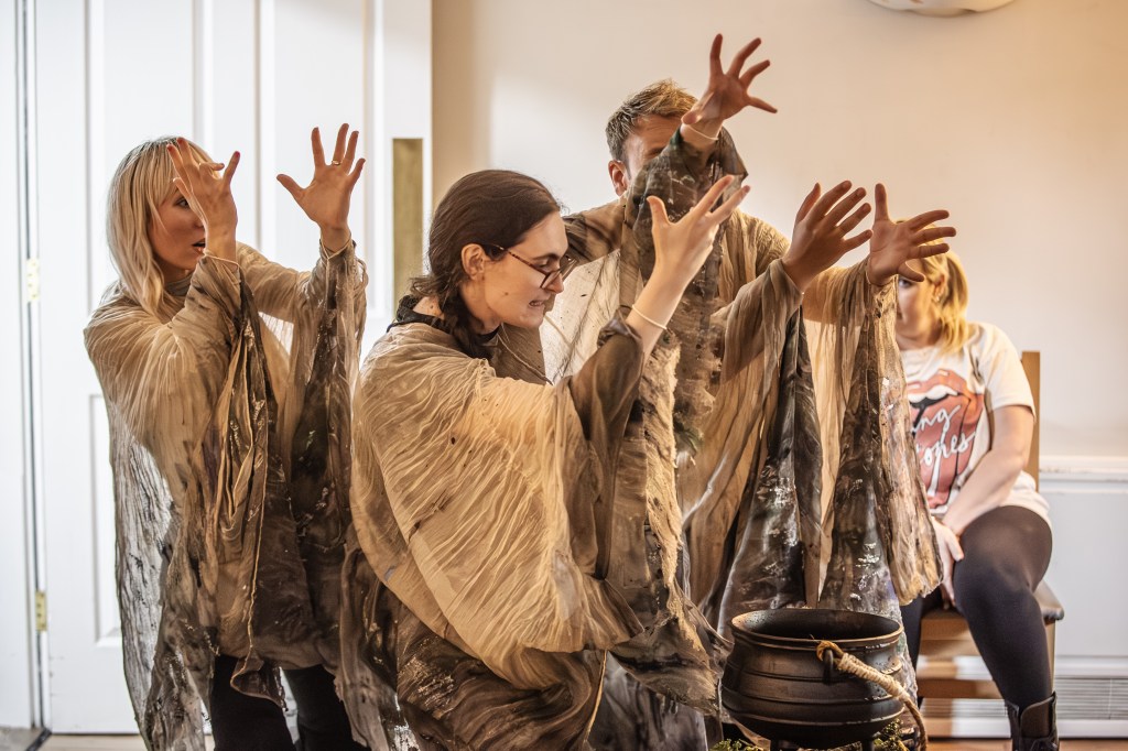 3 white people, dressed in witchy sand coloured robes raise their hands mid spell