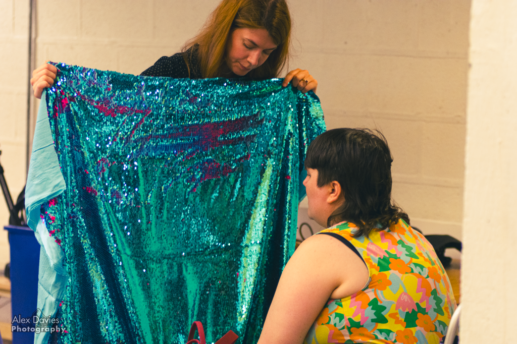 A white woman holds up a blue sequined piece of fabric while Phoebe looks at it