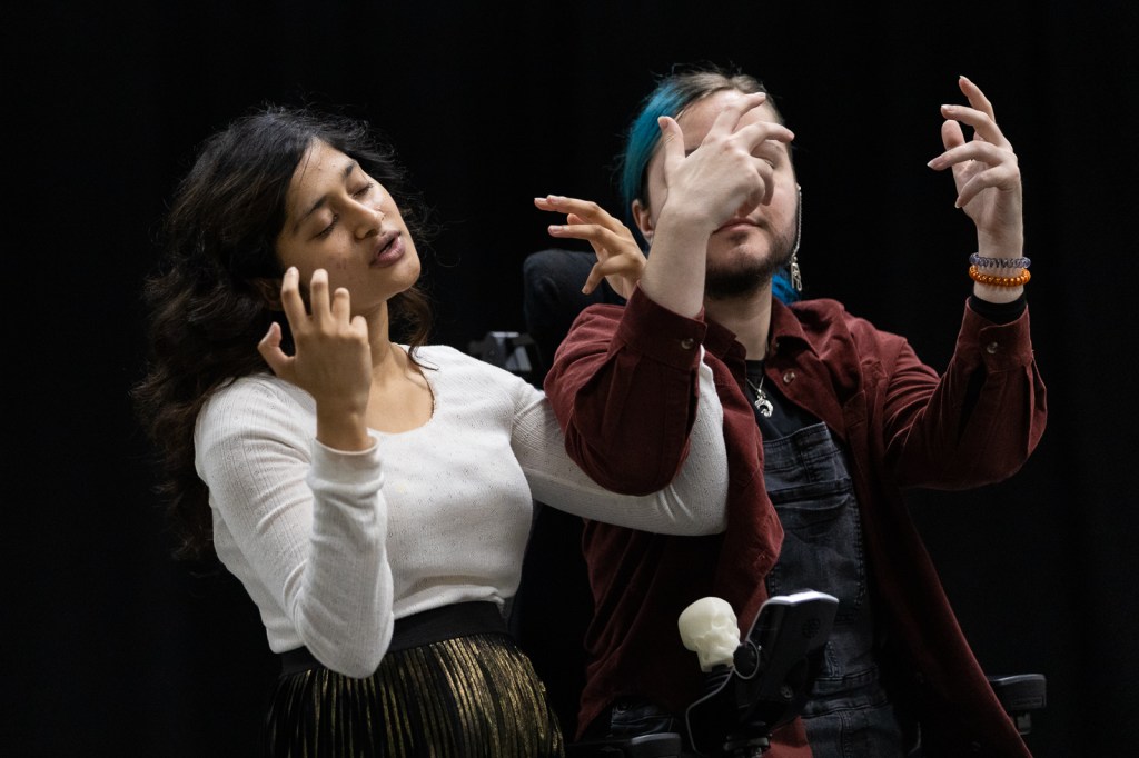A woman of Asian descent stands next to a white non-binary wheelchair user, their arms are intertwined mid BSL signing.